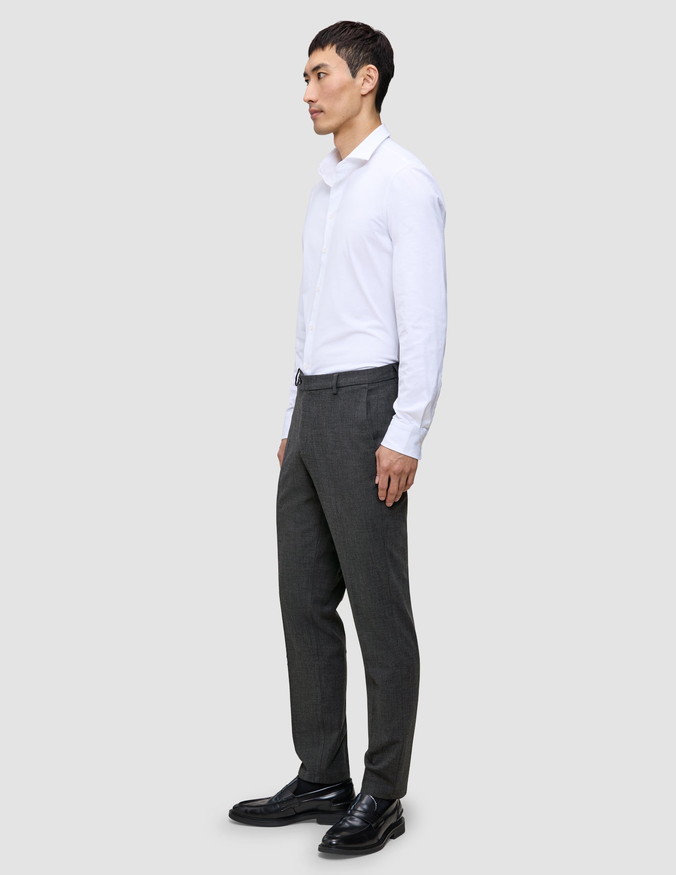 En man klädd i en vit skjorta, byxor av modellen Motion Suit Pants Regular Pepper Grey och svarta loafers i en ren skandinavisk design.